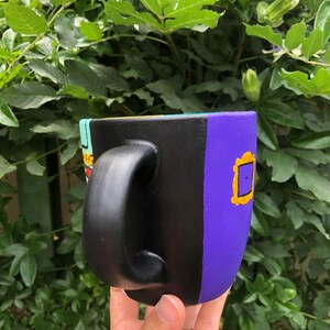 Peut inclure: Une tasse en c&eacute;ramique noire avec un motif violet, jaune et bleu. La tasse a une poign&eacute;e noire et pr&eacute;sente un cadre jaune avec un int&eacute;rieur violet.