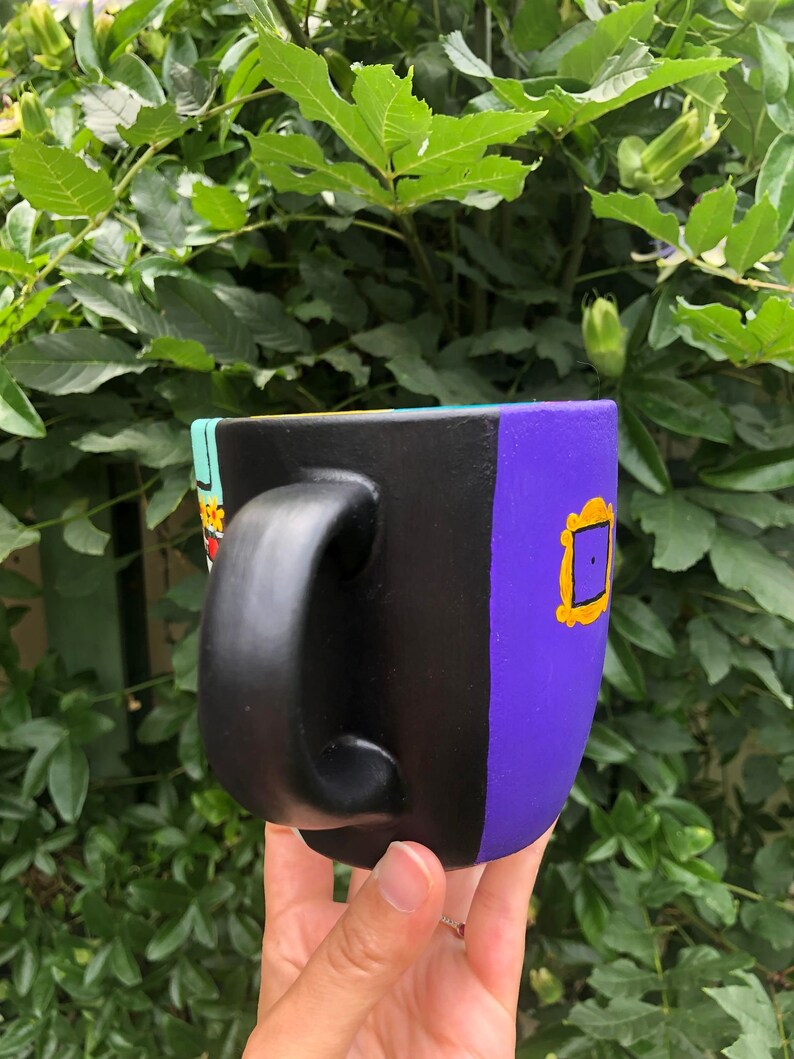 Peut inclure: Une tasse en c&eacute;ramique noire avec un motif violet, jaune et bleu. La tasse a une poign&eacute;e noire et pr&eacute;sente un cadre jaune avec un int&eacute;rieur violet.
