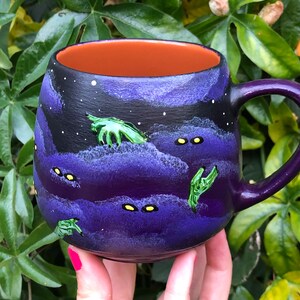 K&ouml;nnte beinhalten: Eine Keramiktasse mit lila Griff und Innenraum. Die Au&szlig;enseite zeigt ein dunkelblaues und violettes Nachthimmeldesign mit wei&szlig;en Sternen, gr&uuml;nen H&auml;nden und gelben Augen. Die Tasse wird in einer Hand mit pinkfarbenem Nagellack gehalten.