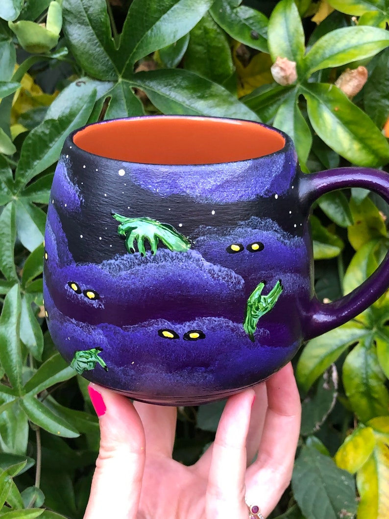 K&ouml;nnte beinhalten: Eine Keramiktasse mit lila Griff und Innenraum. Die Au&szlig;enseite zeigt ein dunkelblaues und violettes Nachthimmeldesign mit wei&szlig;en Sternen, gr&uuml;nen H&auml;nden und gelben Augen. Die Tasse wird in einer Hand mit pinkfarbenem Nagellack gehalten.