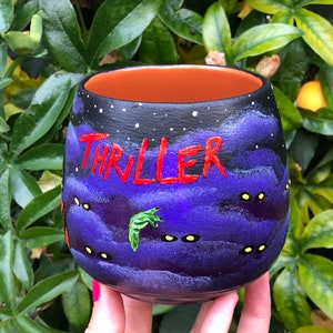 K&ouml;nnte beinhalten: Handbemalter Keramikbecher mit schwarzem Au&szlig;enrand und orangefarbenem Innenraum. Der Becher zeigt das Wort "THRILLER" in Rot, ein lila-schwarzes Nachthimmel-Design und gelb leuchtende Augen. Eine gr&uuml;ne Hand ist ebenfalls auf den Becher gemalt.