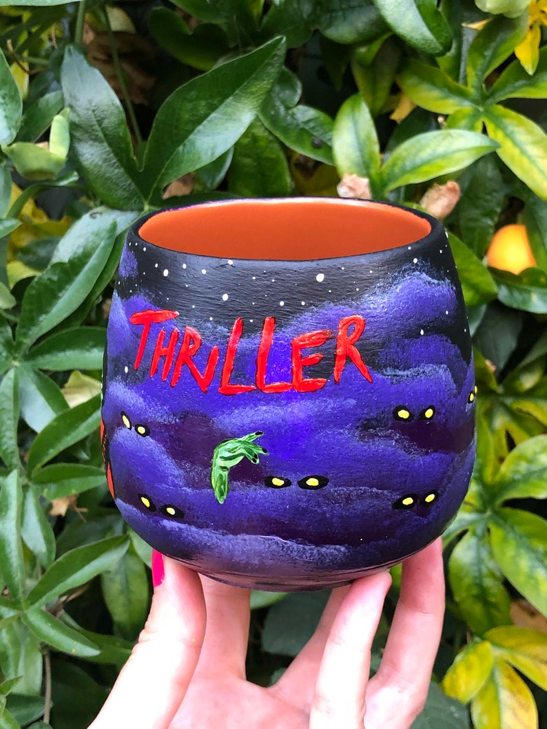K&ouml;nnte beinhalten: Handbemalter Keramikbecher mit schwarzem Au&szlig;enrand und orangefarbenem Innenraum. Der Becher zeigt das Wort "THRILLER" in Rot, ein lila-schwarzes Nachthimmel-Design und gelb leuchtende Augen. Eine gr&uuml;ne Hand ist ebenfalls auf den Becher gemalt.