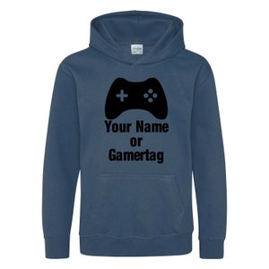 以下が含まれることがあります： 黒いビデオゲームコントローラーのグラフィックと「Your Name or Gamertag」というテキストが書かれた濃い青色のフード付きスウェットシャツです。