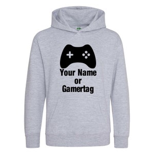 以下が含まれることがあります： グレーのフード付きスウェットシャツに、黒いゲームパッドのグラフィックと「Your Name or Gamertag」のテキストがプリントされています。