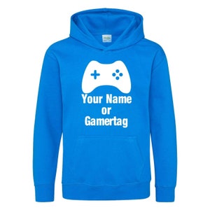 以下が含まれることがあります： 白いビデオゲームコントローラーのグラフィックと「Your Name or Gamertag」というテキストが書かれた明るい青色のフード付きスウェットシャツです。