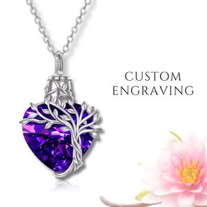 Peut inclure: Un collier argenté avec un pendentif en cristal violet foncé en forme de cœur. Un motif d'arbre est superposé au cristal. Les mots "CUSTOM ENGRAVING" sont imprimés à droite. Une fleur rose est en bas à droite.