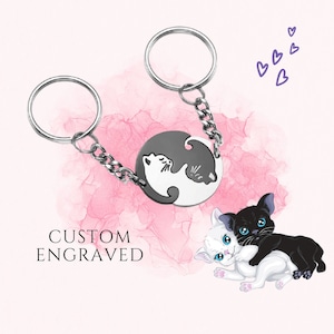 May include: A silver keychain with two interlocking circles featuring black and white cat silhouettes. The keychain is attached to a silver chain. The text "CUSTOM ENGRAVED" is visible in the image.
