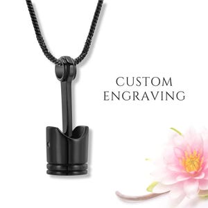 Op de afbeelding: Een zwarte ketting met een hanger in de vorm van een zuiger. De hanger hangt aan een zwarte ketting. De woorden "CUSTOM ENGRAVING" zijn zichtbaar. Een roze bloem staat in de rechter benedenhoek.