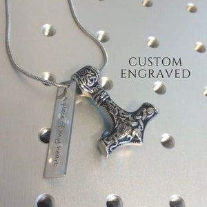Op de afbeelding: Een zilveren ketting met een Mjolnir-hanger en een zilveren tag gegraveerd met de tekst "A piece of my heart". De hanger heeft ingewikkelde details en een getextureerd oppervlak. De ketting ligt op een witte achtergrond met de tekst "CUSTOM ENGRAVED" eroverheen.