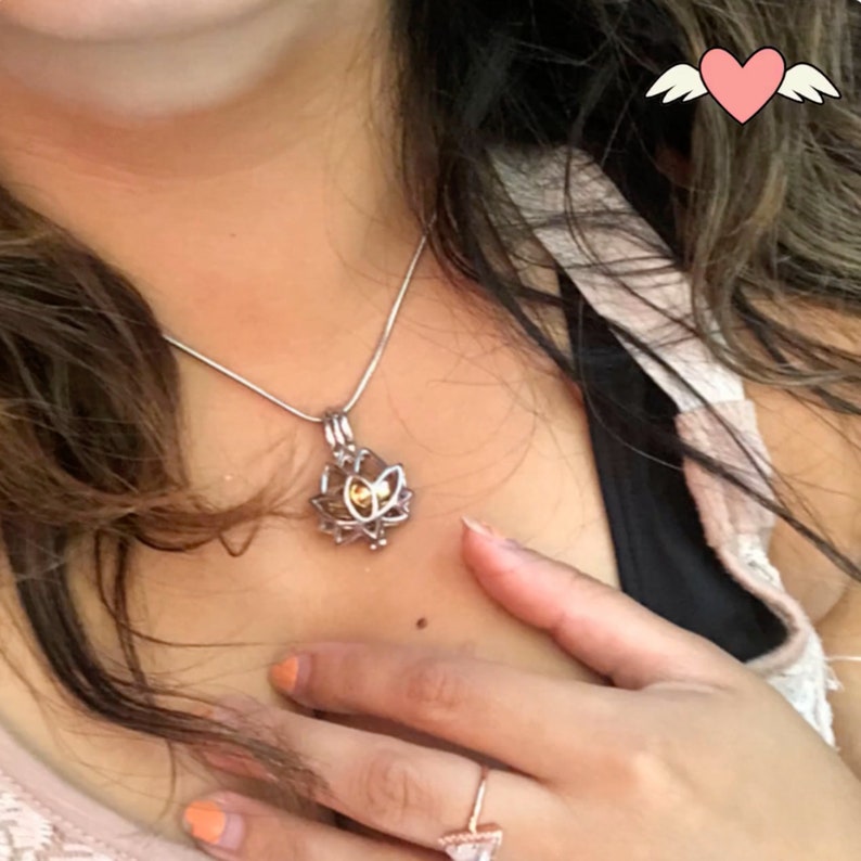 May include: A silver-colored necklace with a lotus flower pendant. The pendant has a cage-like design with a yellow center. The necklace is worn on a person's neck.