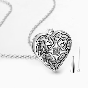 Puede incluir: Un collar con un dije en forma de corazón plateado con un intrincado diseño floral y un pequeño girasol en el centro. El dije se abre y viene con un pequeño embudo y una herramienta para insertar objetos.