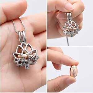 May include: A silver-colored lotus flower pendant necklace with a rose gold-colored inner compartment. The pendant is shown in multiple views, including a close-up of the small, cylindrical urn with the word "DAD" engraved on it.