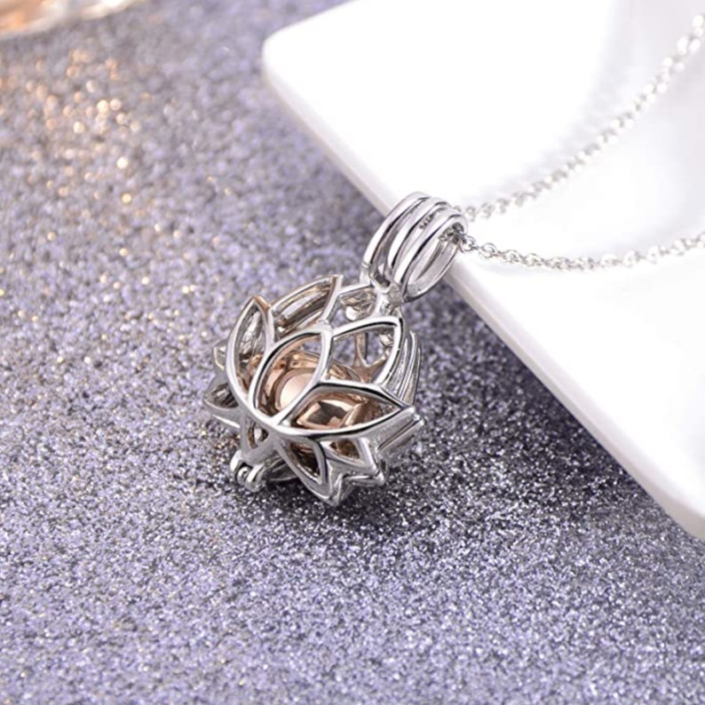 May include: Silver-colored pendant necklace featuring a lotus flower design. The pendant has an openwork design with a central, light-colored sphere. The necklace is displayed on a glittery, silver surface.
