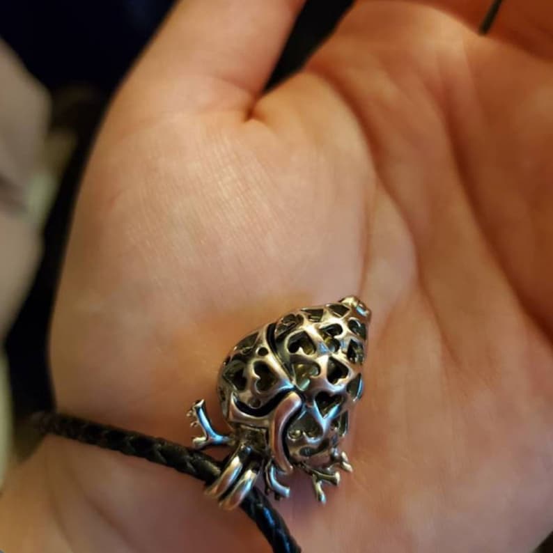 May include: A silver heart-shaped charm with intricate cutouts hangs from a black leather cord. The charm is a popular style for aromatherapy necklaces.