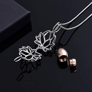 May include: A silver-colored lotus flower pendant necklace with a matching rose gold urn. The pendant is intricately designed with openwork petals. The necklace is displayed on a reflective black surface.
