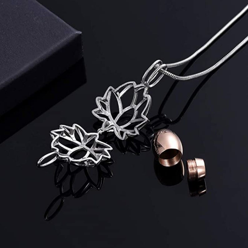 May include: A silver-colored lotus flower pendant necklace with a matching rose gold urn. The pendant is intricately designed with openwork petals. The necklace is displayed on a reflective black surface.