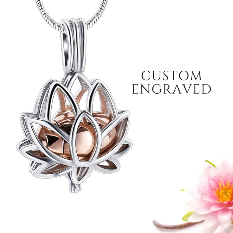 May include: Silver-tone lotus flower pendant necklace with a rose gold interior. The pendant is suspended from a delicate chain. The text "CUSTOM ENGRAVED" is displayed next to the pendant. A pink flower is in the background.