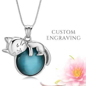 Puede incluir: Un collar con colgante de gato plateado con un orbe de piedra preciosa azul. El gato está acurrucado y el collar tiene una cadena delicada. El texto "CUSTOM ENGRAVING" es visible.