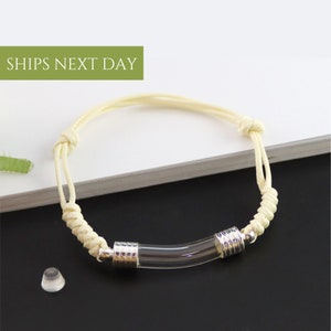 May include: A white cord bracelet with a clear glass tube charm. The tube is accented with silver metal rings.