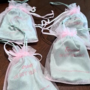 May include: Four small, light blue drawstring bags with pink tulle. The bags have the text "Forever in my heart" printed on them in pink. The text "AndForeverInMyHeart.com" is printed on one of the bags.