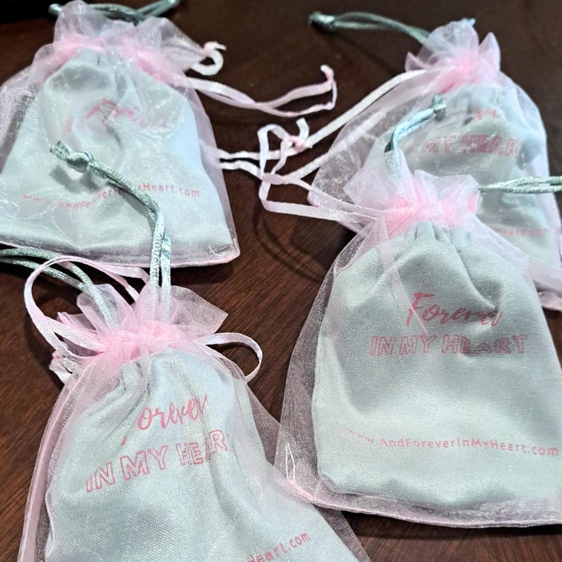 May include: Four small, light blue drawstring bags with pink tulle. The bags have the text "Forever in my heart" printed on them in pink. The text "AndForeverInMyHeart.com" is printed on one of the bags.