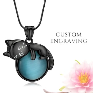 Può includere: Collana con ciondolo a forma di gatto nero con una sfera di pietra preziosa blu. Il gatto è sdraiato, con dettagli bianchi sul viso. La collana ha una catena nera. Il testo "CUSTOM ENGRAVING" è visibile.