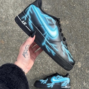 Könnte beinhalten: Schwarze und blaue Custom-Sneaker mit einem Blitz-Design. Die Sneaker haben ein weißes Swoosh-Logo und das Wort "AIR" an der Seite.