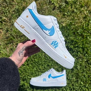 Peut inclure: Baskets blanches avec des détails bleus, dont un logo et un symbole d'équipe. Les chaussures présentent des détails dorés en forme d'étoile et de texte, avec "AIR" imprimé sur le côté. Les baskets sont posées sur un fond d'herbe verte.