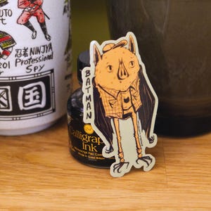 May include: A sticker of a bat character wearing a hat and a plaid shirt, with the word "Batman" printed vertically. The sticker is next to a black ink bottle and a white mug with Japanese text and illustrations.
