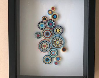 Puede incluir: Obra de arte enmarcada con un diseño de papel enrollado de círculos concéntricos en tonos de azul, amarillo, rojo y negro.