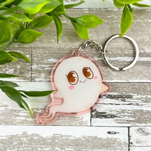 Może przedstawiać: Brelok do kluczy w kształcie różowego i białego ducha z uroczą kreskówkową twarzą i dużymi oczami. Duch jest przezroczysty i ma srebrny kółko do kluczy.