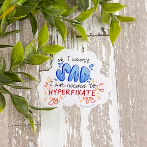 May include: A white sticker with the text "oh I wasn't SAD I just needed to HYPERFIXATE" in blue and red. The sticker is cloud-shaped with a light blue outline and is decorated with stars and rectangles.