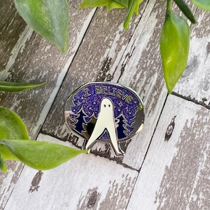 Pode incluir: Um broche de esmalte prateado com um fantasma branco em frente a um céu noturno estrelado roxo e azul. O broche diz "Eu Acredito" em um contorno prateado ao redor do fantasma.