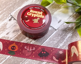 Puede incluir: Un rollo de cinta washi roja con un acento de lámina dorada. La cinta presenta un patrón repetitivo de lindos críptidos de dibujos animados, que incluyen un fantasma, una polilla, un ciervo, una linterna de calabaza y un sasquatch. La cinta está etiquetada "10m washi tape Round Cryptids @lespritdesigns | www.lespritdesigns.com"