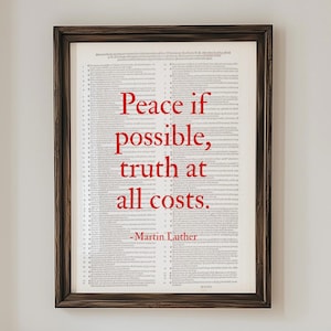 May include: A framed print with the quote "Peace if possible, truth at all costs." in red text. The print is in a dark wood frame and hangs above a wooden console table. Two white ceramic vases and a wooden bowl are on the table.