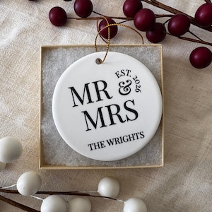 May include: A round white ceramic ornament featuring black text that reads MR & MRS, EST. 2026, and THE WRIGHTS. The ornament has a gold hanging string and is presented inside a square cardboard box with white padding, surrounded by berry sprigs.