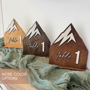May include: Three wooden mountain-shaped table numbers in varying shades of brown. Each has a white mountain peak design and the words "table 1" in a cursive font. The table numbers are displayed on a sage green fabric.