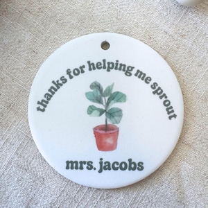 May include: A round white ceramic ornament featuring a small green potted plant illustration in the center. The text "thanks for helping me sprout" is printed in a dark green font above the plant, and "mrs. jacobs" is printed below it.
