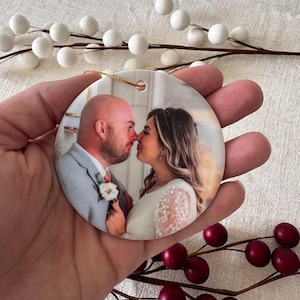 May include: A round ceramic ornament featuring a printed photograph of a couple in wedding attire. The man wears a light gray suit with a white boutonniere, and the woman wears a white dress with lace sleeves. A gold string is attached to the top.