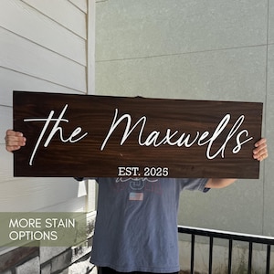 May include: A rectangular dark brown wooden sign featuring white raised lettering that reads The Maxwells in a cursive script, with the text EST. 2025 printed in a simple white sans-serif font centered below the main name on the board.