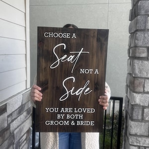 May include: A dark brown wooden sign with white lettering. The sign reads "CHOOSE A SEAT NOT A SIDE YOU ARE LOVED BY BOTH GROOM & BRIDE". The sign is rectangular and appears to be handmade.