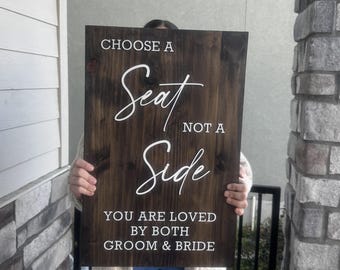 May include: A dark brown wooden sign with white lettering. The sign reads "CHOOSE A SEAT NOT A SIDE YOU ARE LOVED BY BOTH GROOM & BRIDE". The sign is rectangular and appears to be handmade.