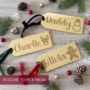 May include: Three rectangular gold-toned acrylic gift tags with black, green, and red ribbons. Each tag features a custom name and a holiday icon: Daddy with a snowman, Charlie with a reindeer, and Olivia with a gingerbread man. Text reads 15 ICONS TO PICK FROM.