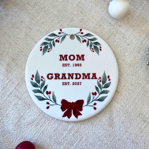 May include: A round white ceramic ornament featuring a green watercolor leaf wreath with red berries and a red bow at the bottom. The text reads MOM EST. 1985 and GRANDMA EST. 2027 in dark red lettering. A small hole is at the top for hanging.