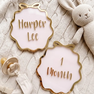 May include: Two scalloped-edge acrylic plaques with gold trim and a bow detail at the top. One plaque features the name Harper Lee in gold script, and the other displays the text 1 Month in gold script. A beige pacifier and a knitted toy are present.