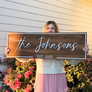 May include: A wooden sign with the text "the Johnsons" in white cursive lettering. Below the name, the text "EST. 2025" is displayed. The sign has a white border and a dark wood grain effect. The sign is rectangular.