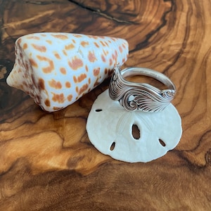 May include: A silver spoon ring with an ornate design, resting on a white sand dollar. The ring is engraved with the words "11.00 Gold Filled".