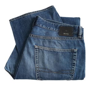 Pode incluir: Jeans azuis dobrados com um design clássico de cinco bolsos. Os jeans apresentam um patch de couro marrom escuro com a marca "BOSS" nas costas. O denim tem uma lavagem média com desbotamento sutil.