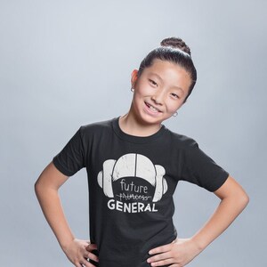May include: A young person wearing a black t-shirt with the text "Future Princess General" in white, paired with blue jeans featuring a star pattern. The person is also wearing black sneakers with white laces.