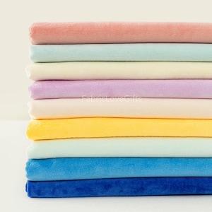 May include: A stack of colourful plush fabric rolls in shades of pink, blue, yellow, and purple. The fabric has a soft, velvety texture. The image is well-lit, showcasing the vibrant colours and the texture of the material.